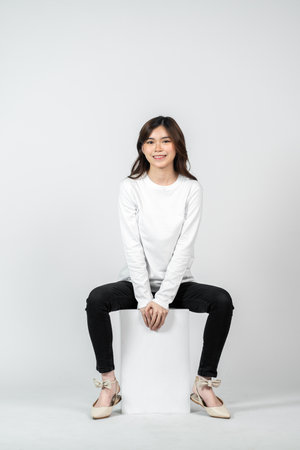 A young asian woman wearing a white blank long sleeve shirt doing a pose while sitting, isolated on the white backgroundの写真素材