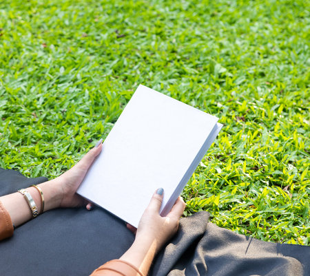 Image of a woman's hand holding out a white blank hardcover book with the grass as the background, hardcover mockup template imageの写真素材