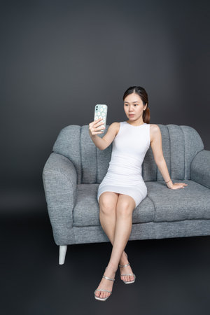 Portrait of a stunning Asian woman taking a selfie on a stylish sofa with an alluring pose, shirt dress mockup template imageの写真素材