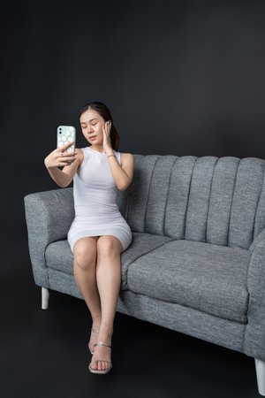 Indoor portrait of an alluring Asian woman capturing her beauty in a selfie on a luxurious sofa, shirt dress mockup template imageの写真素材