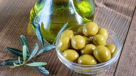 Canned green olives in glass transparent bowl and olive oil bottle on the wooden table of the kitchen. Healthy diet food. の写真素材