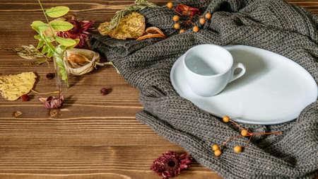White cup and saucer on the warm winter pillover, autumnal colorful leaves and dry flowers on the wooden background. Copy space for your text.の写真素材