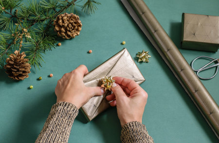 Female hands packing present box. Christmas tree branch, cones and wrapping paper on the green...