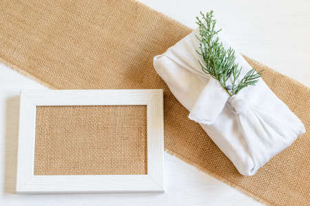 Alternative handmade eco friendly gift for Christmas or any other holiday.Gift box wrapped white clothes in Japanese Furoshiki style, picture frame with natural burlap blank. Handmade gifts, care packaging, diy concepts.の写真素材