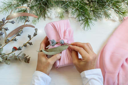 Female hands packaging with love Christmas gift. Handmade gift box wrapping in pink cloth in japanese furoshiki style. Decorated home table, top view.の写真素材