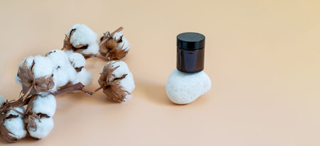 Glass jar with cosmetics on the white stone podiun and natural cotton branch on the beige background. Mockup banner photo with copy space for displaying your natural eco friendly product, your logo or text.の写真素材