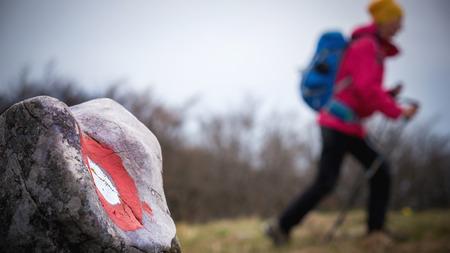 A female hiker hiking in the background with the trail mark on a rock in the foreground in the mountainsの写真素材