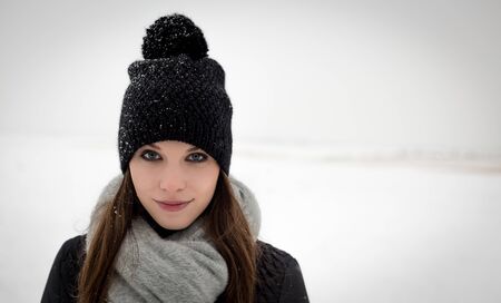 Outdoor portrait of young caucasian girl with long brown hair and blue eyes in winter hat with pom-pom, scarf and out of focus snowy plains in background. Girl has a very decent smile on her face.の写真素材