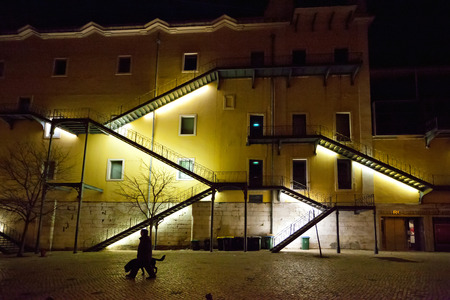 Illuminated fire escapes on the exterior wall of an apartment block at night in a Lisbon street with a person taking their dog for a walk in the foregroundのeditorial素材