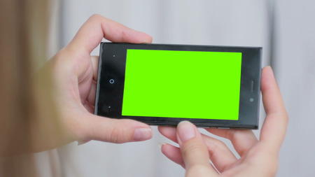 Woman looking at horizontal smartphone with green screen. Close up shot of woman's hands with mobile. White wooden backgroundの写真素材