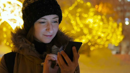 Young pretty woman using smartphone in the city at night. Christmas, winter, technology and holiday conceptの写真素材