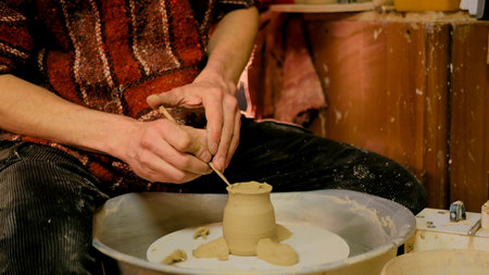 Professional male potter shaping and carving mug with special tool in pottery workshop, studio....