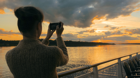 Woman silhouette taking photo of beautiful sunset with smartphone on deck of cruise ship. Sunset light, golden hour. Photography, nature and journey conceptの写真素材