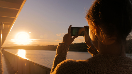 Woman silhouette taking photo of beautiful sunset with smartphone on deck of cruise ship. Sunset light, golden hour. Photography, nature and journey conceptの写真素材