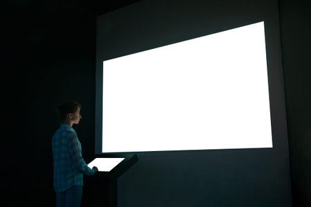 Woman using electronic kiosk and looking at white blank large interactive wall display in dark room...