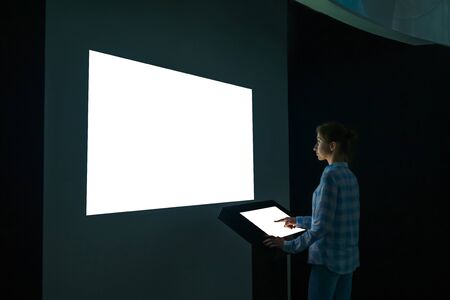 Mock up, futuristic, template and education concept. Woman using electronic kiosk and looking at white blank large interactive wall display in dark room of modern technology museum - mockup imageの写真素材