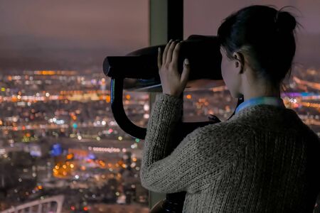 Young woman looking through tourist telescope from skyscraper, exploring night cityscape. Light city street bokeh illumination at evening. Relax, discover and journey conceptの写真素材