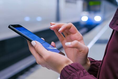 Woman hands using black smartphone on subway platform. Train arriving at station - close up view. Urban and technology conceptの写真素材