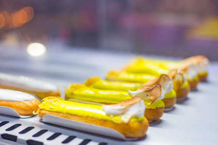 Close up view - assortment of delicious fresh cake eclairs with cream for sale on counter of shop,...