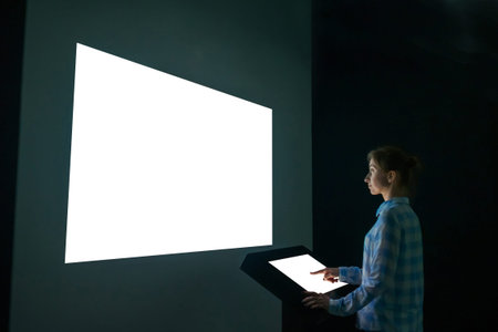 Woman using electronic kiosk and looking at white blank large interactive wall display in dark room...
