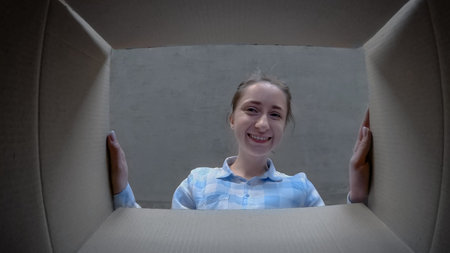 Happy positive woman in plaid shirt opening cardboard box with gift, looking inside and smiling -...