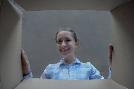 Happy positive woman in plaid shirt opening cardboard box with gift, looking inside and smiling - pov low angle view. Unboxing, unpacking, holiday, birthday, celebration, emotions, surprise conceptの写真素材