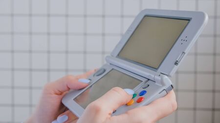 Gaming, hobby, technology, portable, entertainment, video game and leisure time concept. Woman gamer hands using grey handheld game console against tile wall at home - close up side viewの写真素材