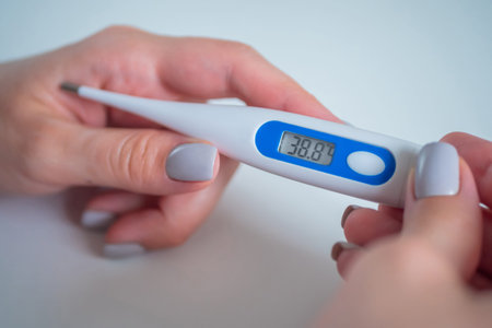 Woman hands holding white digital medical thermometer with high temperature - close up view,...