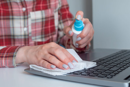 Woman hands spraying antiseptic, cleaning laptop keyboard with disinfectant wet wipe on white table....