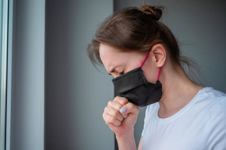 Woman wearing black medical face mask, feeling sick, suffering from coughing, looking out of window in grey room - side view. Self isolation, quarantine, COVID-19, coronavirus, safety conceptの写真素材