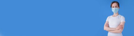 Wide banner copyspace - woman wearing medical face mask, white shirt, looking at camera against blue background at home. Self isolation, prevention, quarantine, COVID19, coronavirus, safety conceptの写真素材