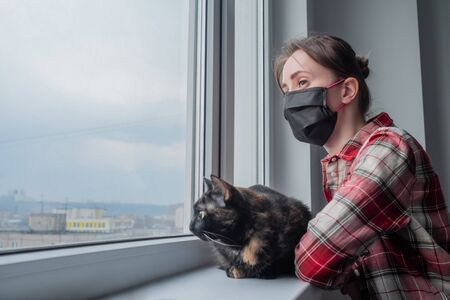 Portrait of pensive woman in medical face mask and black cat looking out of window in grey room at home - side view. Self isolation, prevention, quarantine, COVID 19, coronavirus, safety conceptの写真素材