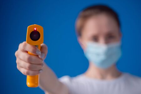 Woman in medical face mask holding yellow pyrometer to measure temperature toward camera - close up view, selective focus. Healthcare, measurement, disease, infection, coronavirus conceptの写真素材