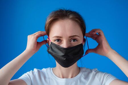 Portrait of woman in white shirt putting on black medical face mask and looking at camera in room with blue wall at home: close up. Self isolation, prevention, quarantine, COVID19, coronavirus conceptの写真素材