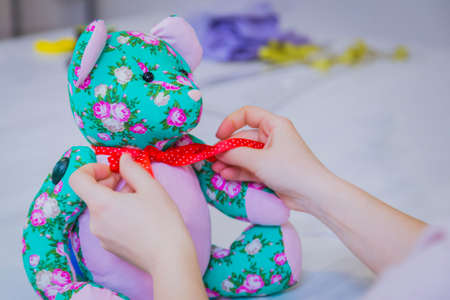Tailor woman, toymaker tying bow on neck of colorful teddy bear at sewing studio - close up view. Handmade, craft and toy making conceptの写真素材