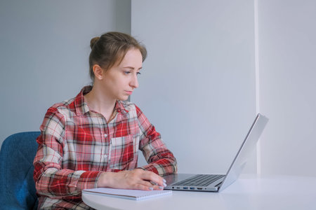 Woman in red plaid shirt using laptop computer, watching webinar, lecture in grey room at home....