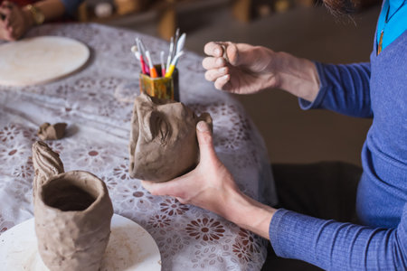 Professional male potter shaping mug in shape of human face with special tool in pottery workshop,...