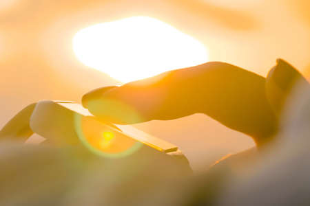 Close up: woman hand using wearable white smartwatch device against warm sunset sky - scrolling and touching. Sun lens flare, golden hour. Relax, leisure time and technology conceptの写真素材