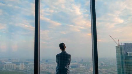 Back view of pensive woman silhouette looking at cityscape through window of skyscraper. Summer...