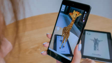 MOSCOW, RUSSIA - AUGUST 1, 2019. Woman using smartphone and tablet with augmented reality application - virtual 3d models of animals - giraffe. Future, AR, nature, education, technology conceptのeditorial素材