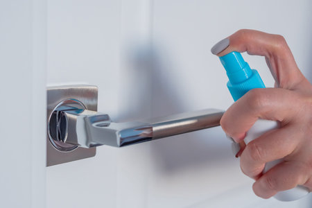 Woman spraying antiseptic and cleaning door handle - close up view. Disinfection, protection,...