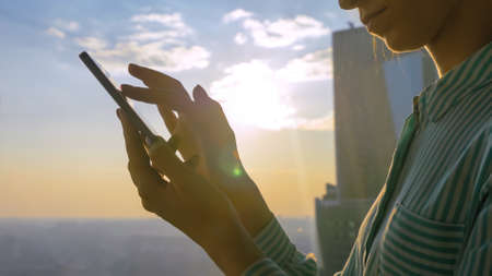 Woman using smartphone device against sunset cityscape view through window of skyscraper: scrolling and touching. Sun lens flare, golden hour. Leisure time, sightseeing, technology conceptの写真素材