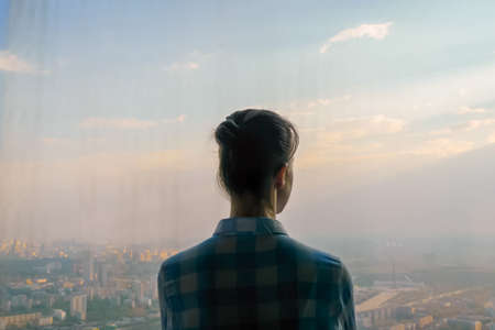 Back view of pensive woman silhouette looking at cityscape through window of skyscraper. Summer time, clear sky, sunset. Success, opportunity, sightseeing, discover and future conceptの写真素材