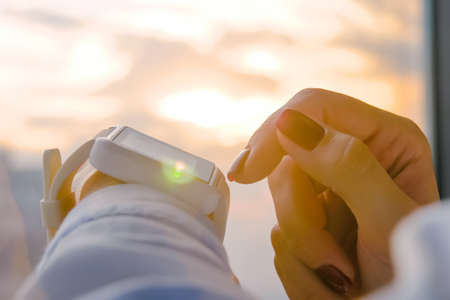 Close up: woman hand using wearable white smartwatch device against warm sunset sky - scrolling and touching. Sun lens flare, golden hour. Relax, leisure time and technology conceptの写真素材