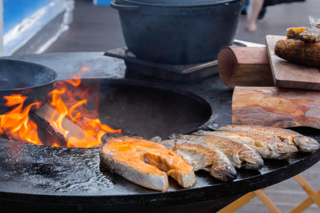 Process of grilling salmon steaks and mackerel fish on black brazier at summer local food market -...