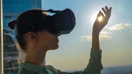 Woman using virtual reality headset and moving hand against view on cityscape from panoramic skyscraper window in office. Sun lens flare. VR, sightseeing, discover, technology conceptの写真素材