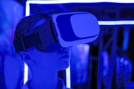 VR, augmented reality, immersive, entertainment concept. Woman using virtual reality headset and looking right at interactive technology exhibition with blue projector light illuminationの写真素材