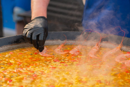 Chef hand cooking yellow paella with shrimp, mussel, rice, tomato, spice, saffron in huge paella pan at summer food market - close up. Spanish cuisine, seafood, gastronomy, street food conceptの写真素材