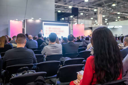 Back view of woman in red dress and unrecognizable people, audience listening lecture, presentation at trade show, conference hall. Business, congress, seminar, corporate, training, education conceptの写真素材
