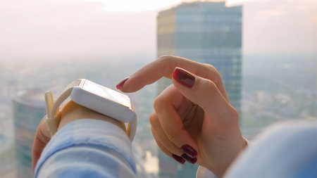 Woman hand using wearable white smartwatch device against view on cityscape from window in office -...